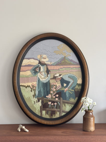Framed needlepoint of two people in a pastoral scene on a wooden shelf.