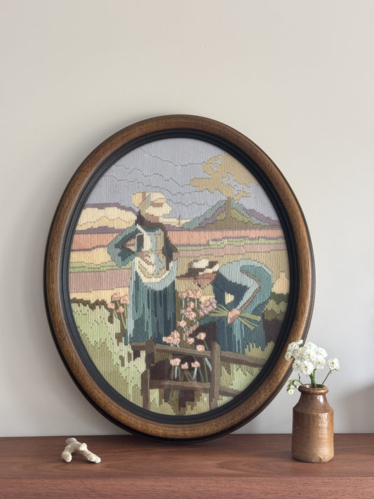 Framed needlepoint of two people in a pastoral scene on a wooden shelf.