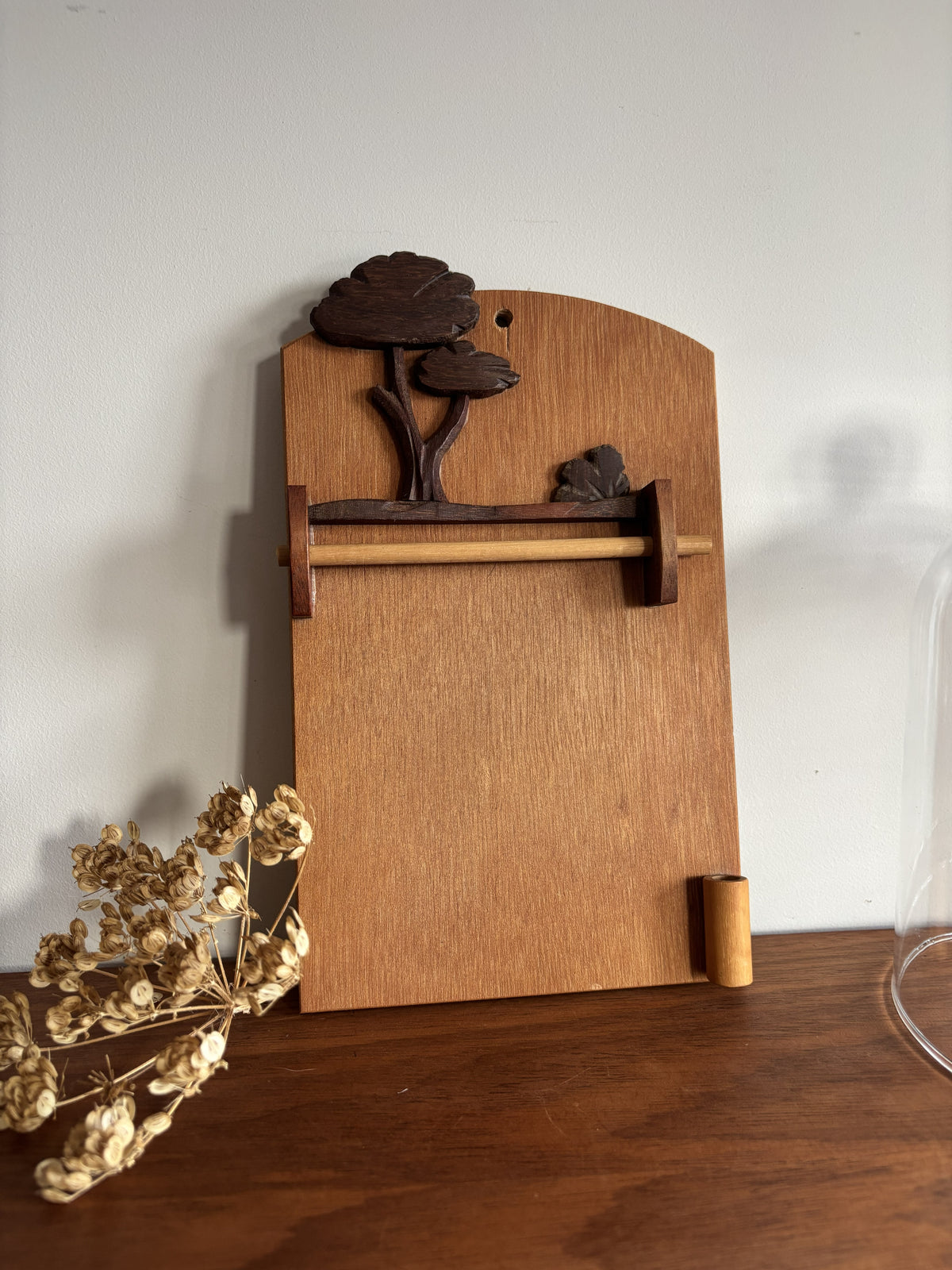 A handcrafted wooden wall-mounted holder with a natural wood grain finish, featuring a decorative carved tree motif at the top and a horizontal wooden bar. The holder is shown standing on a dark wooden surface against a plain white wall, with dried flowers in the foreground.