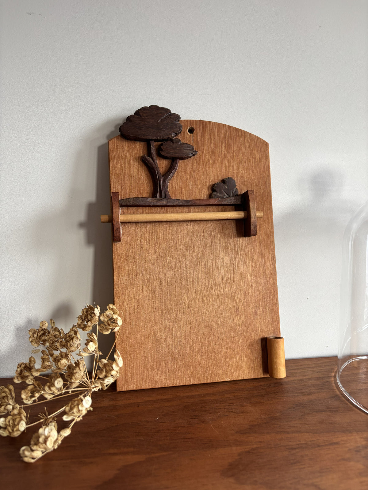 A wooden wall-mounted towel bar with a decorative carved tree at the top, shown resting on a wooden table against a plain light wall with dried flowers nearby.
