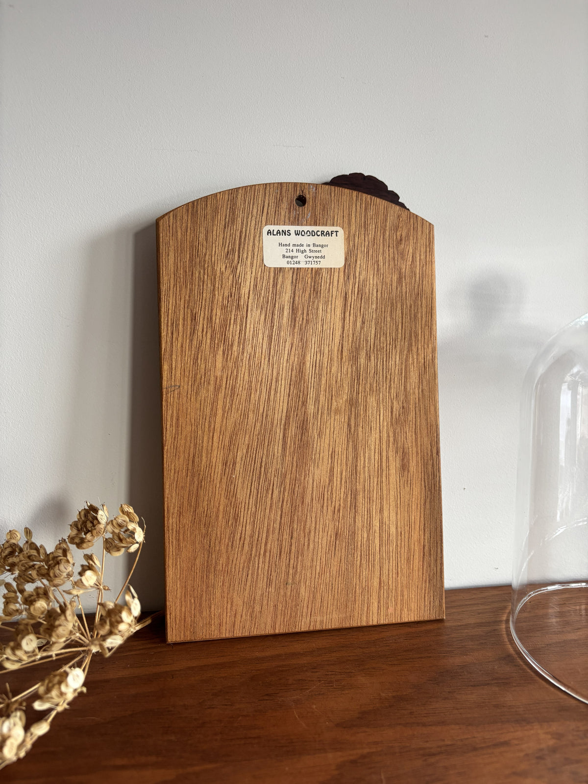 A vertical wood slab with a curved top edge and a small white label reading 'ALANS WOODCRAFT' stands on a wooden surface against a light gray wall. Dried flowers appear at the lower left, and part of a glass dome is visible at the lower right.