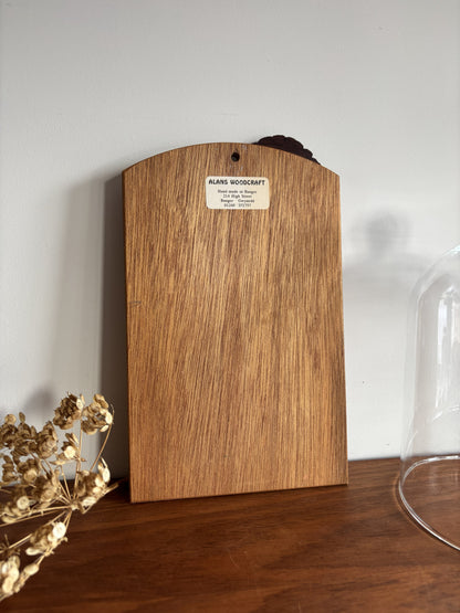 A vertical wood slab with a curved top edge and a small white label reading 'ALANS WOODCRAFT' stands on a wooden surface against a light gray wall. Dried flowers appear at the lower left, and part of a glass dome is visible at the lower right.