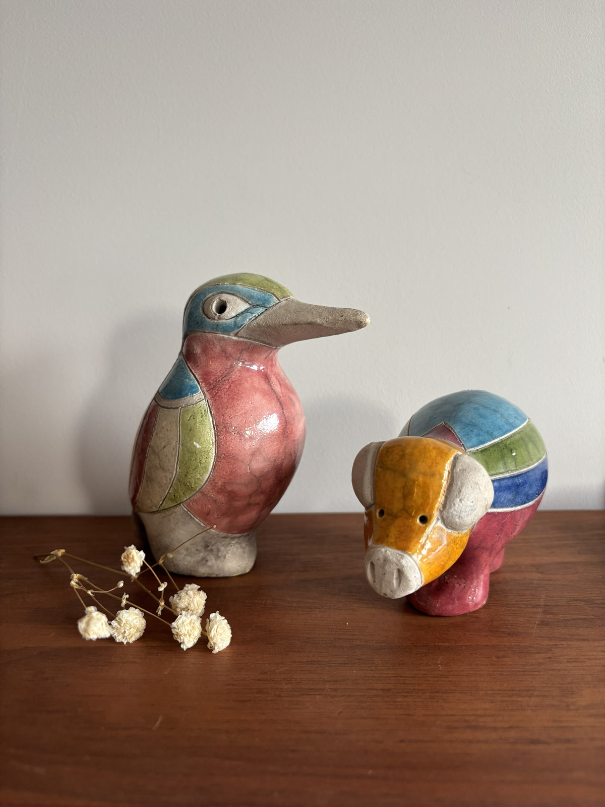 Two colorful glazed ceramic animal figurines, a bird and a pig, displayed on a wooden surface against a plain light gray wall.
