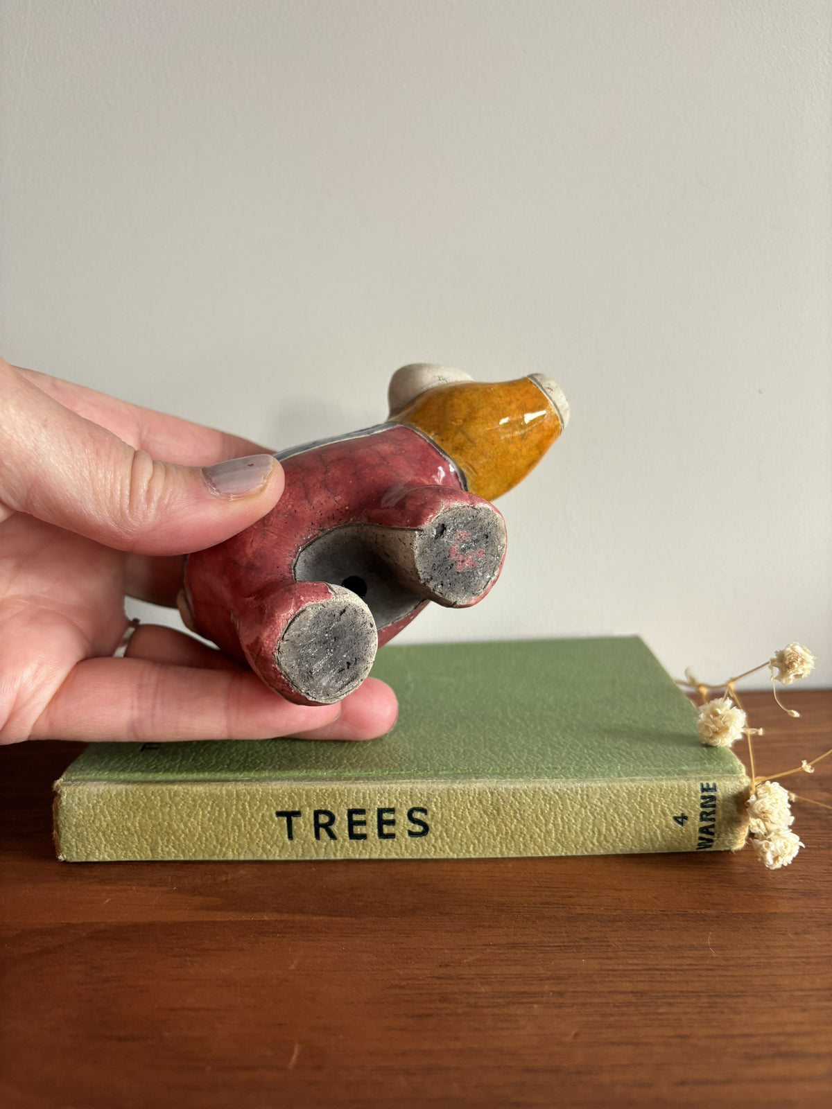 A hand holds a small ceramic pig figurine with a red body and yellow head over a green book on a wooden table.