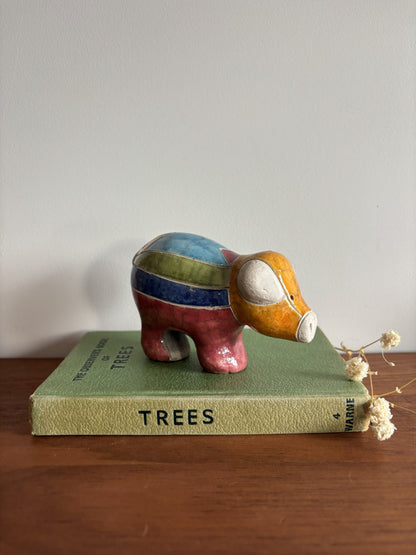 A small hand-painted ceramic pig figurine with colorful stripes in blue, green, red, and orange, standing on top of a vintage green book titled 'TREES' on a wooden surface.