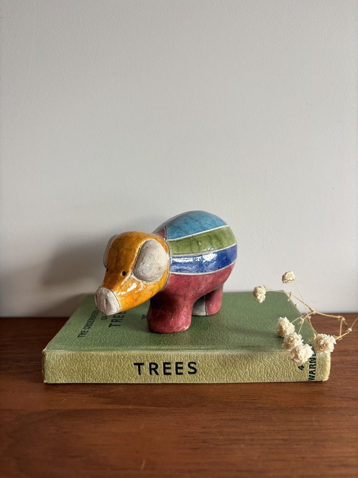 A small ceramic pig figurine with a multicolor striped pattern in orange, yellow, green, blue, and red, displayed on top of a vintage green book titled 'TREES' on a wooden surface against a plain light gray wall.