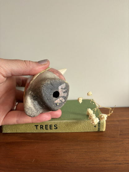 A hand holds a small rustic ceramic bird figurine with a dark gray glazed finish and a visible hole, resting above a green book on a wooden surface.