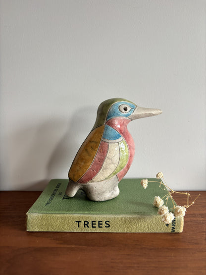 A colorful ceramic bird figurine with painted blue, pink, green, and brown sections, displayed on top of a vintage green book titled 'TREES' on a wooden surface against a plain light gray wall.