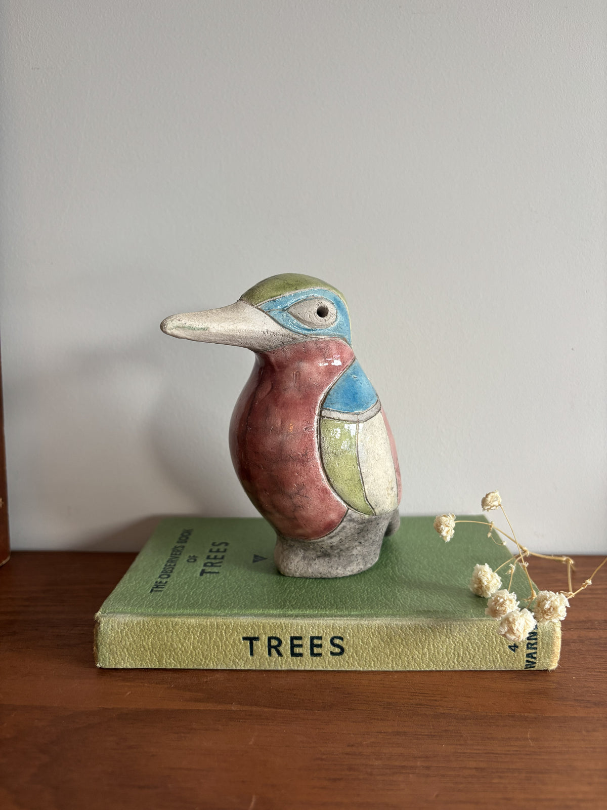 A small ceramic bird figurine with a multicolor glaze sits on top of a vintage green book on a wooden surface against a plain wall.